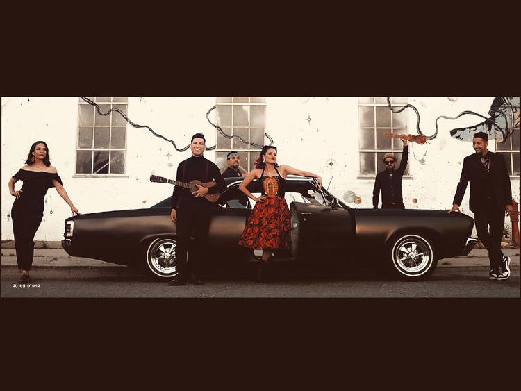 Members of a band hold instruments while standing around a black car